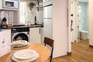 A compact kitchen with a microwave, sink, and washing machine, a dining area with a table set for two, and a view into a bathroom with a toilet and shower. - Pink Calas Blancas Apartment (Torrevieja)