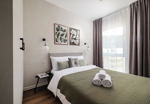 A neatly made bed with a green blanket, two pillows, and rolled towels on the side. There are two wall-mounted light fixtures and framed artwork above the bed. - Wola Redutowa Superior Apartment (Warszawa)