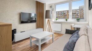 Ein modernes Wohnzimmer mit Flachbildfernseher, einem weißen Sofa, einem Couchtisch, einer Lampe und Blick auf eine Stadtlandschaft durch ein großes Fenster.