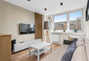 A modern living room with a flat-screen TV, a white sofa, a coffee table, a lamp, and a view of a cityscape through a large window. - Sosnowiec Modern Apartment (Sosnowiec)