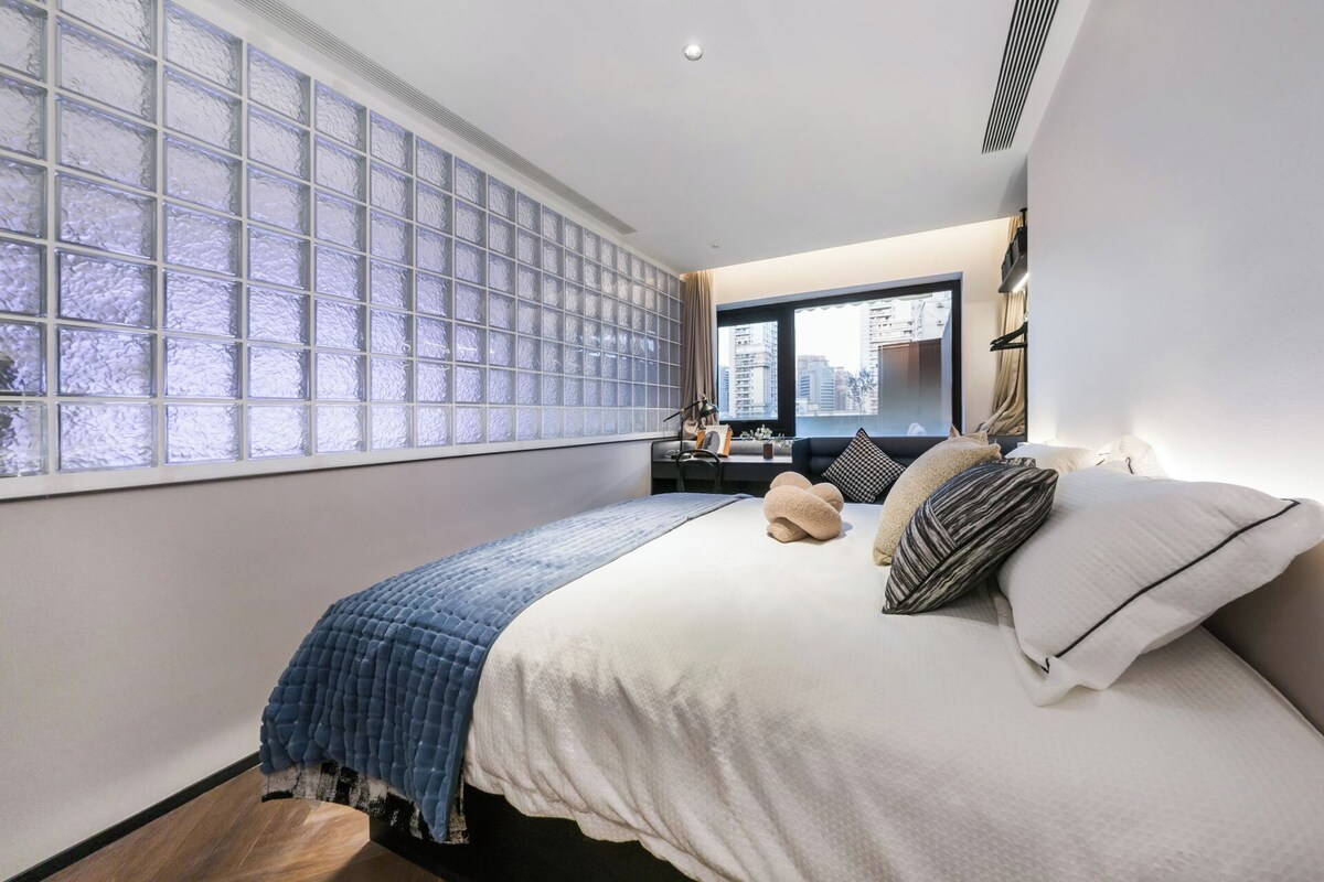 A bedroom with a large window, a bed with pillows, a desk with a chair, and a view of the city.