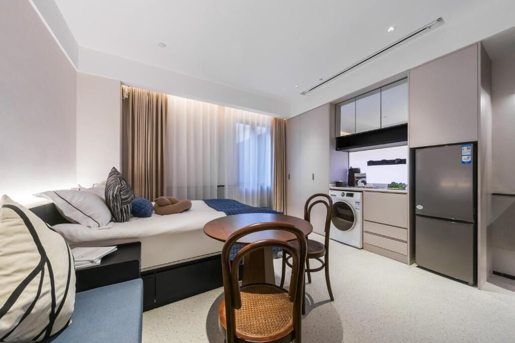 A modern hotel room with a bed, a dining table with chairs, a kitchenette, and a washing machine.