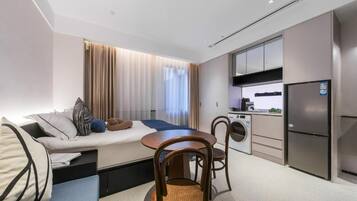 A modern hotel room with a bed, a dining table with chairs, a kitchenette, and a washing machine.