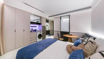 A modern bedroom with a large bed, a built-in washing machine, a small kitchen area, and a seating area with a round table.