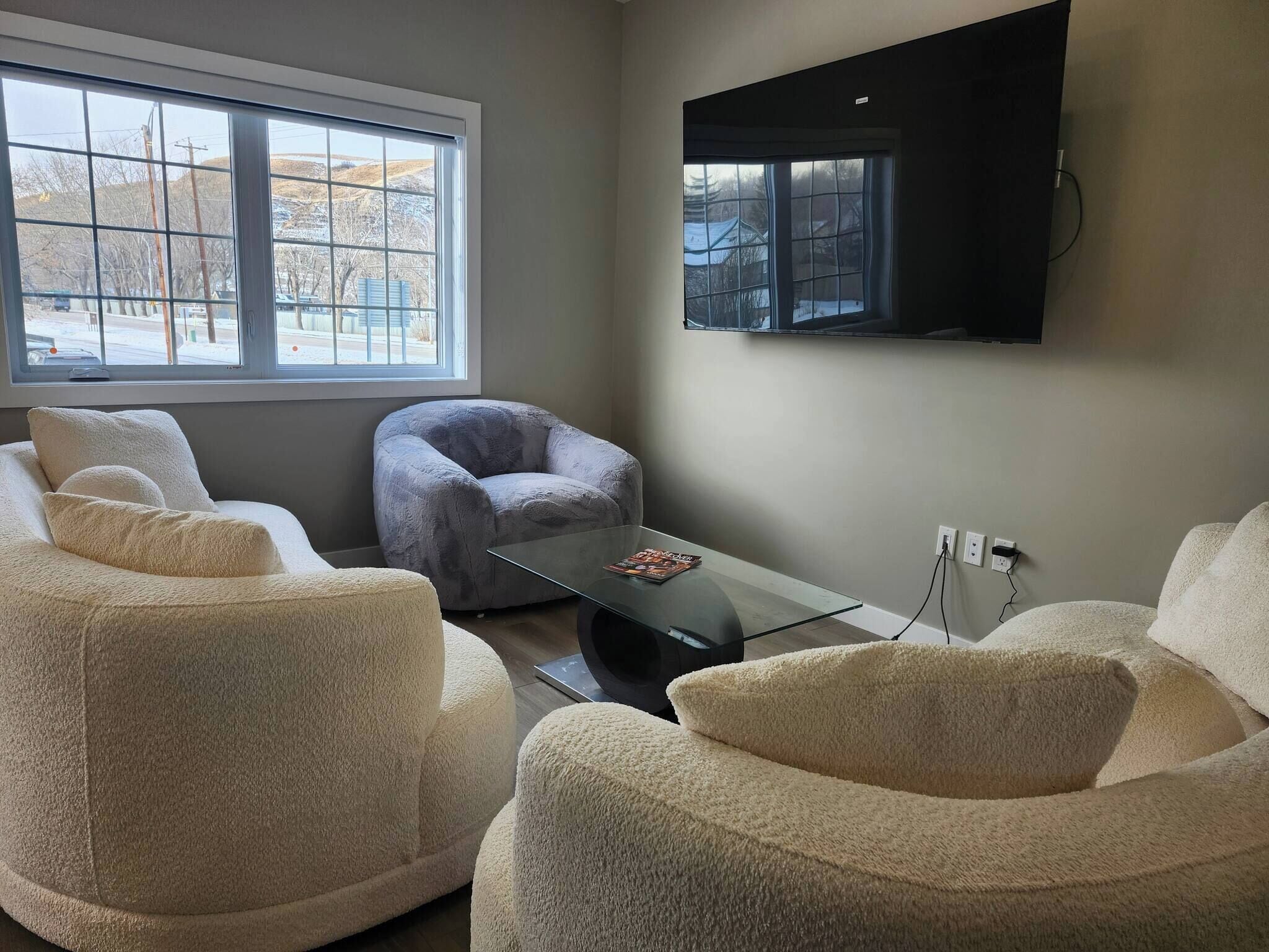 Un salon doté d’une grande fenêtre, d’un fauteuil gris, d’une table basse en verre et d’un téléviseur à écran plat fixé au mur.