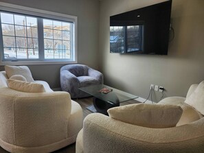A modern living room with a large window, a flat-screen TV mounted on the wall, and plush, textured sofas. - Amazing Medium & Long Term Rentals in Drumheller (Drumheller)