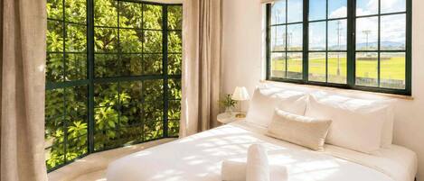 A hotel room with a large bed, a window with a view of greenery, and a nightstand with a lamp.