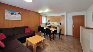 Uma sala moderna com sofá, mesa de centro e área de jantar com cadeiras. Uma cozinha ao fundo, com armários e bancada.