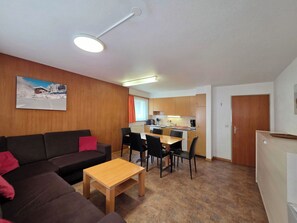 Ein modernes Wohnzimmer mit einer Couch, einem Couchtisch und einem Essbereich mit Stühlen. Im Hintergrund befindet sich eine Küche mit Schränken und einer Arbeitsplatte.
