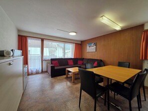 A compact living space with a sofa, a dining table, and chairs, a kitchenette, and a window with curtains. - Am Biel 2 by Interhome (Saas-Fee)
