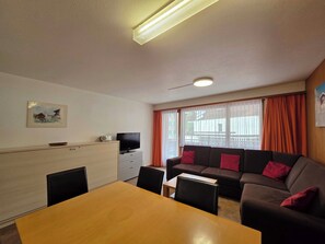 Uma sala de estar mobiliada com sofá, televisão, mesa de jantar e janela com cortinas.