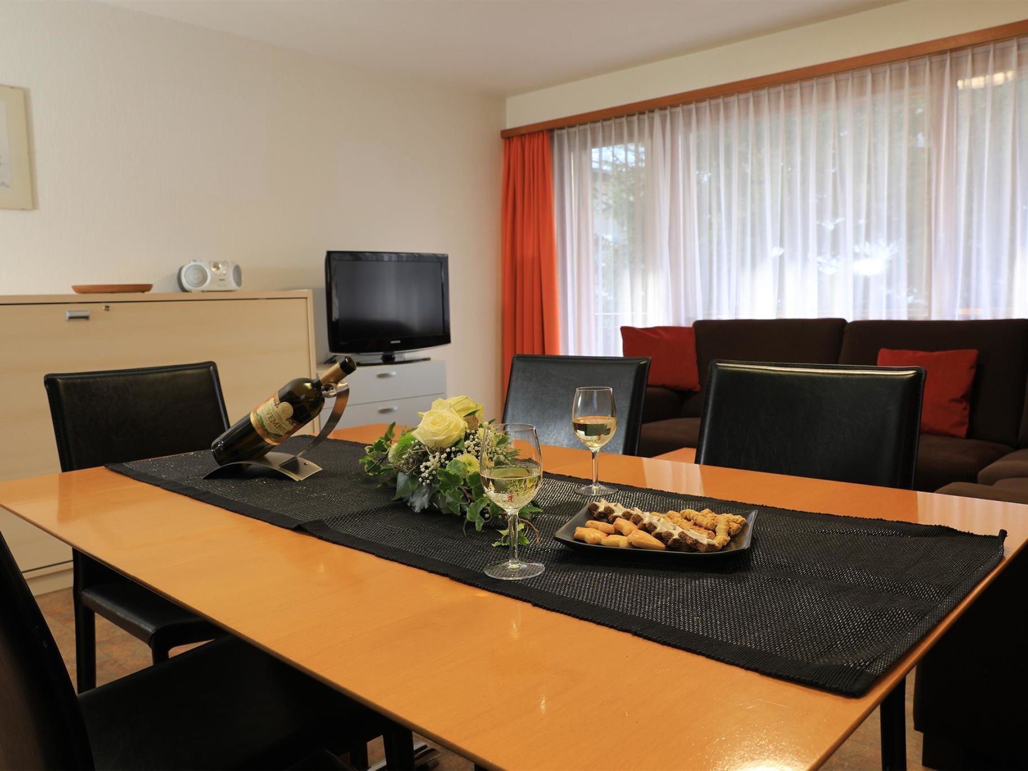 A dining table with a bottle, glasses, and a plate of food in a room with a television and a sofa.