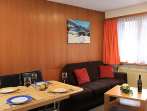 Quarto mobiliado com uma mesa de jantar posta para dois, um sofá com almofadas vermelhas, uma janela com cortinas laranja e um quadro na parede.