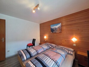 A bedroom with a wooden wall panel, a bed with striped bedding, a chair, and two wall-mounted lights. - Am Biel 3 by Interhome (Saas-Fee)