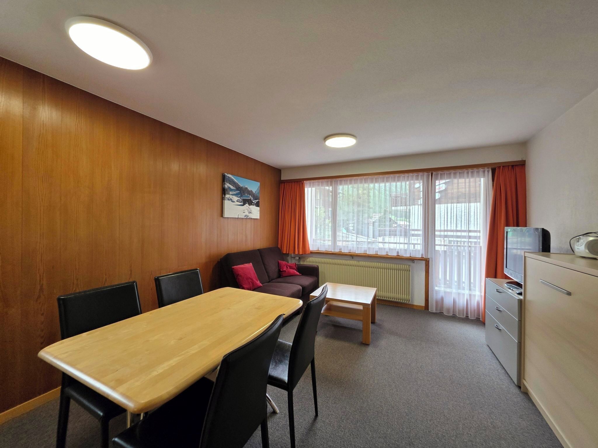 A room with a wooden wall, a dining table with chairs, a sofa, a radiator, and a window with curtains.