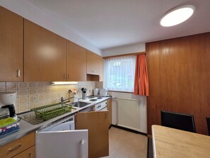 A compact kitchen with wooden cabinets, a built-in oven, a sink, and a window with curtains. - Am Biel 3 by Interhome (Saas-Fee)