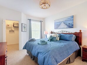 Ein Schlafzimmer mit Bett, Nachttisch, Lampe, einem Fenster mit Vorhängen, einer Tür und einem Gemälde an der Wand.