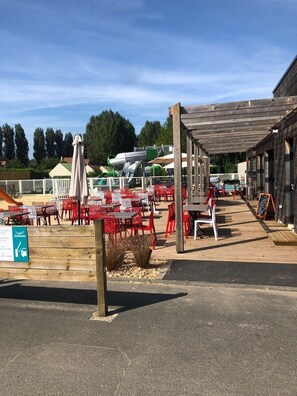 An outdoor seating area with tables and chairs, a covered walkway, and a building with a wooden facade. - Mobile home with a patio and the campground's pool (Ouistreham)