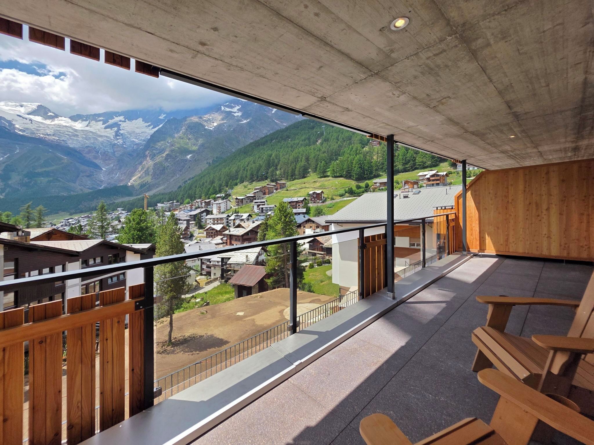 Een balkon met uitzicht op een bergachtig dorp, houten meubels en een betonnen plafond.