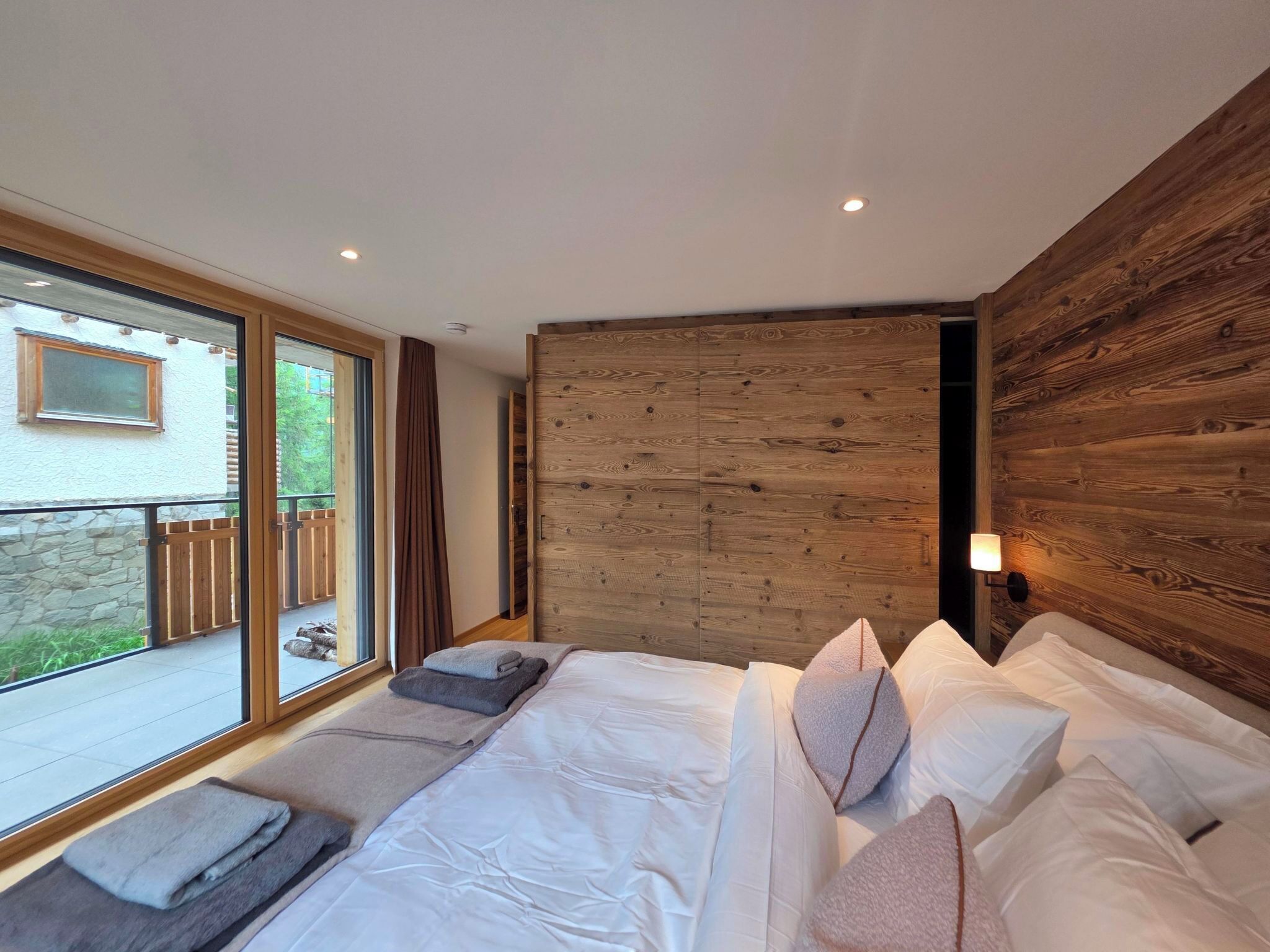 A modern bedroom with a large bed, a wooden headboard, and a view of the outdoors through a sliding door.