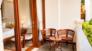 A hotel room with a bed, a bedside table with a lamp, a wooden chair, a small round table, and a curtain.