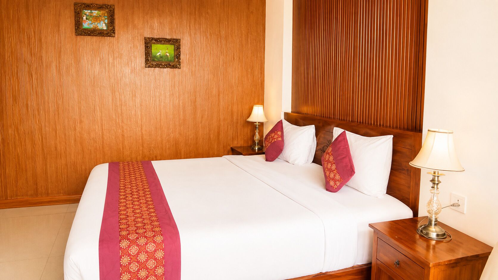 Quarto de hotel com cabeceira de madeira, cama com roupa de cama branca e detalhes vermelhos, duas luminárias de apoio e duas fotos emolduradas na parede.