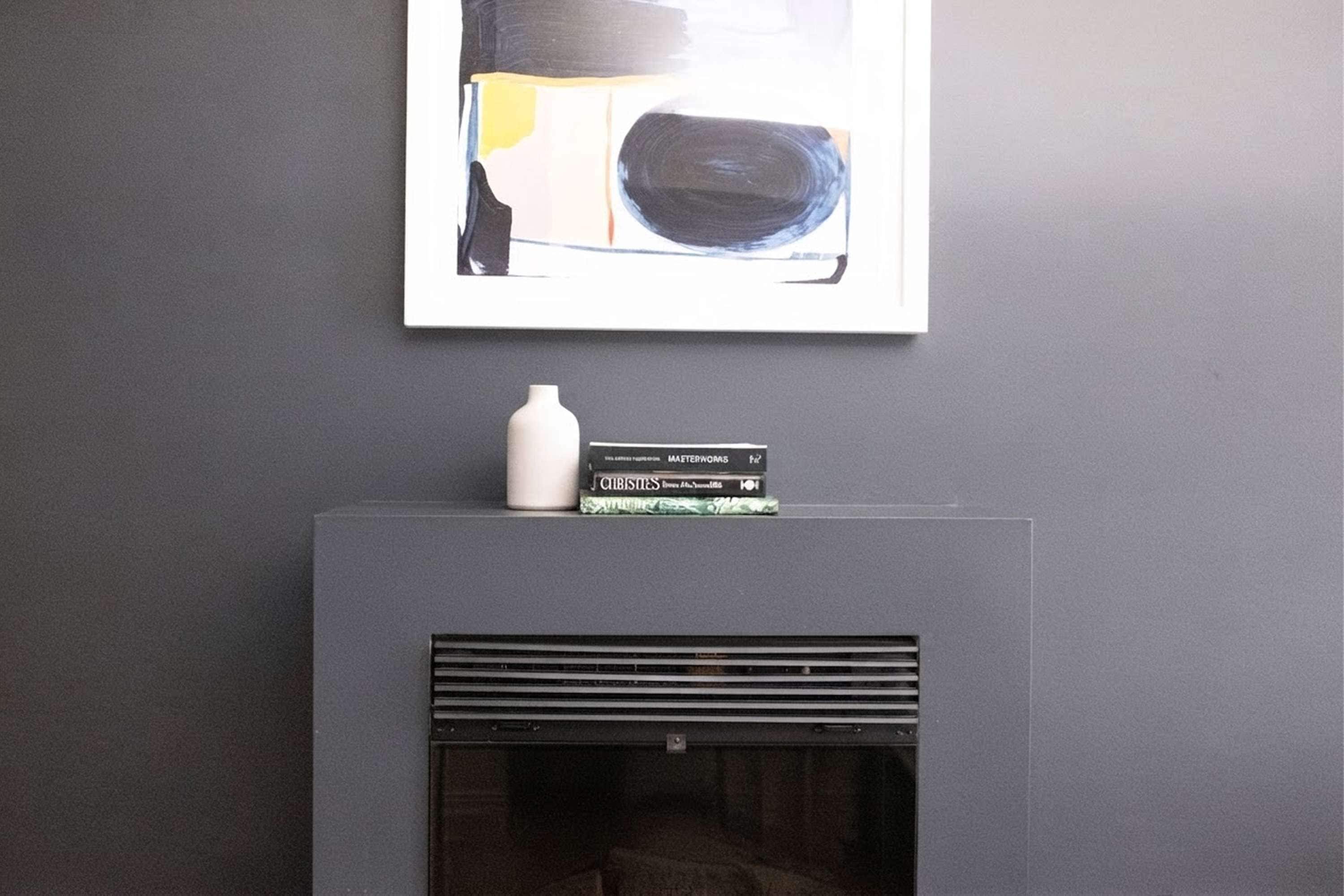 A fireplace with a modern design, a framed abstract painting above it, and a small white vase on top of a stack of books.