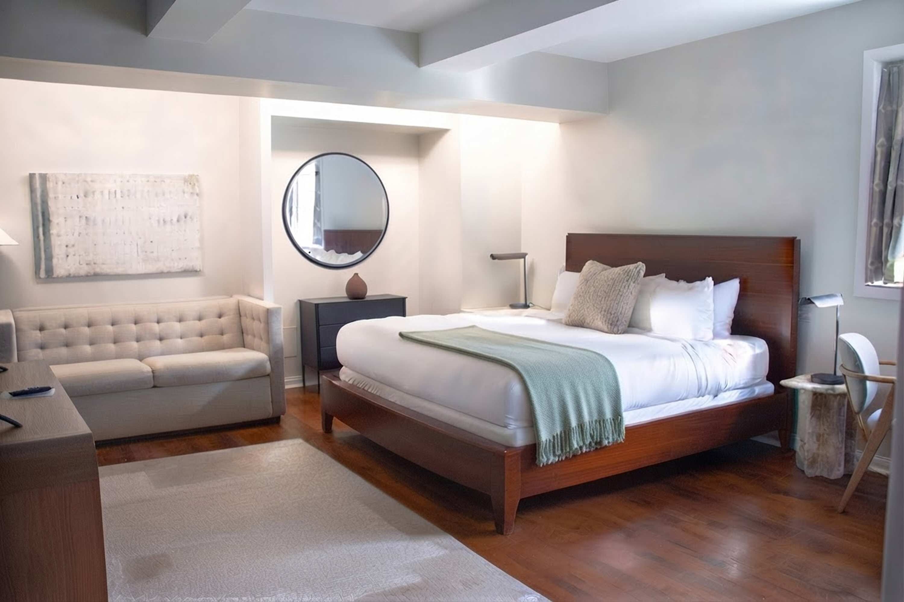A bedroom with a bed, a sofa, a nightstand, a mirror, and a wall-mounted artwork.