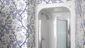 A bathroom with a shower enclosure, a white bathtub, and floral wallpaper. - Queen Double Room | StayCozy at The Arco (Philadelphia)