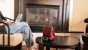 A room with a fireplace, a sofa, a chair, a table, and a red handbag. - Queen Double Room | StayCozy at The Arco (Philadelphia)