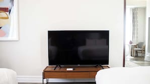 A modern living room with a flat-screen TV on a wooden stand, a white sofa, and a small table with a remote control. - Queen Double Room | StayCozy at The Arco (Philadelphia)