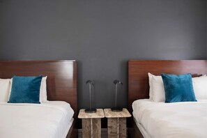A hotel room with two beds, each with a wooden headboard and blue pillows. There are two small tables with lamps between the beds. - Queen Double Room | StayCozy at The Arco (Philadelphia)