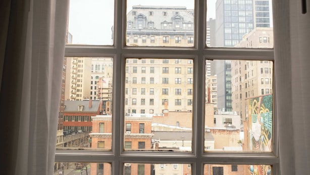 A view through a window with multiple panes, displaying a cityscape with various buildings, including a prominent brick building and a modern high-rise. - Queen Double Room | StayCozy at The Arco (Philadelphia)