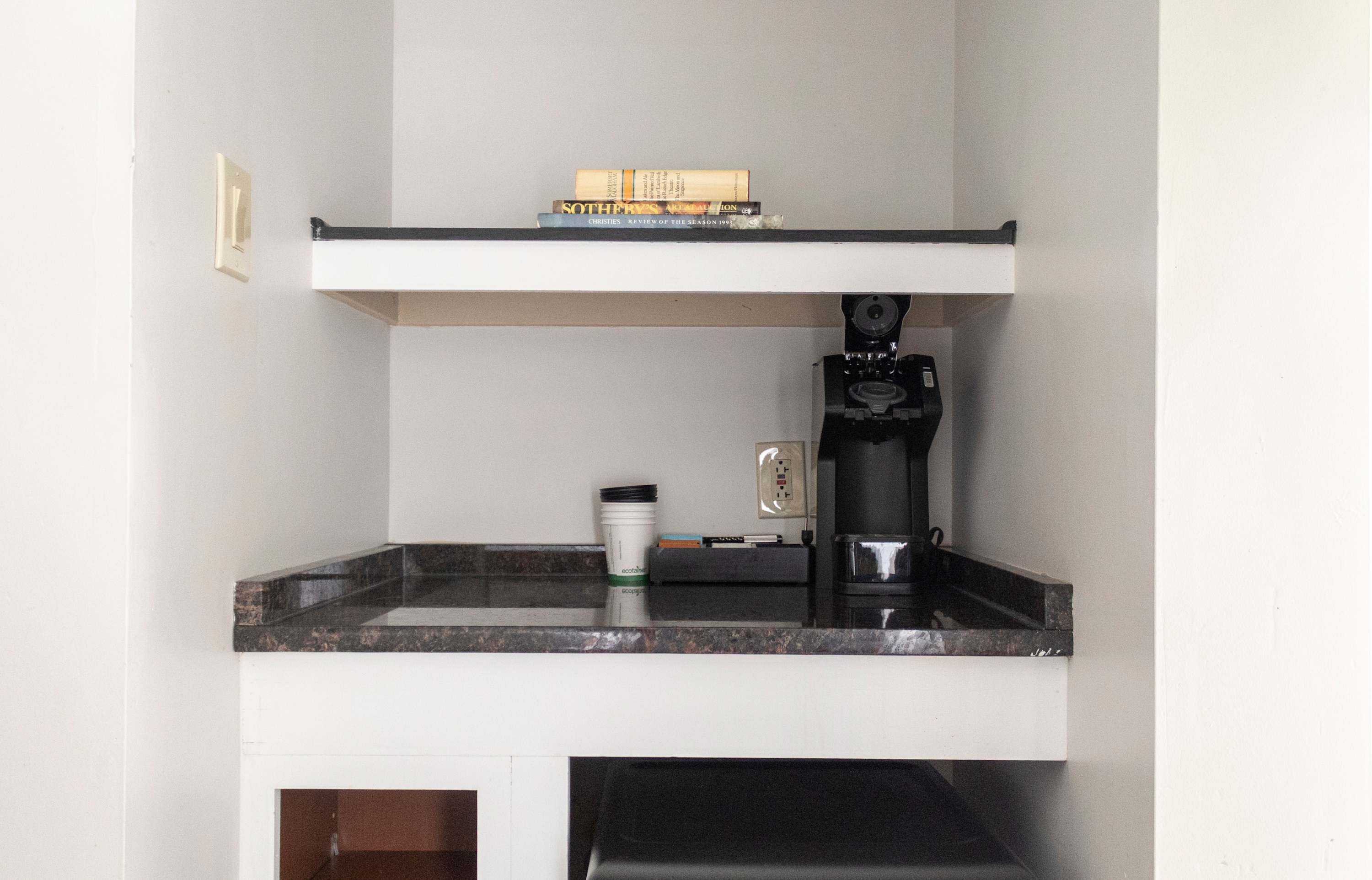 Une cuisine équipée d’un plan de travail, d’une machine à café, d’une prise électrique et d’une pile de livres sur une étagère.