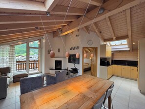 Een ruime woonkamer met een houten eettafel, grijze banken en een aan de muur bevestigde televisie.