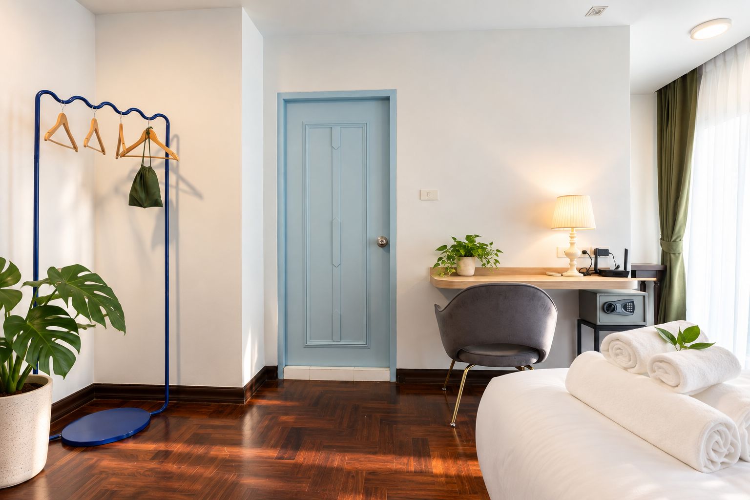 A hotel room with a bed, a desk with a lamp, a chair, a blue door, and a coat rack with hangers.
