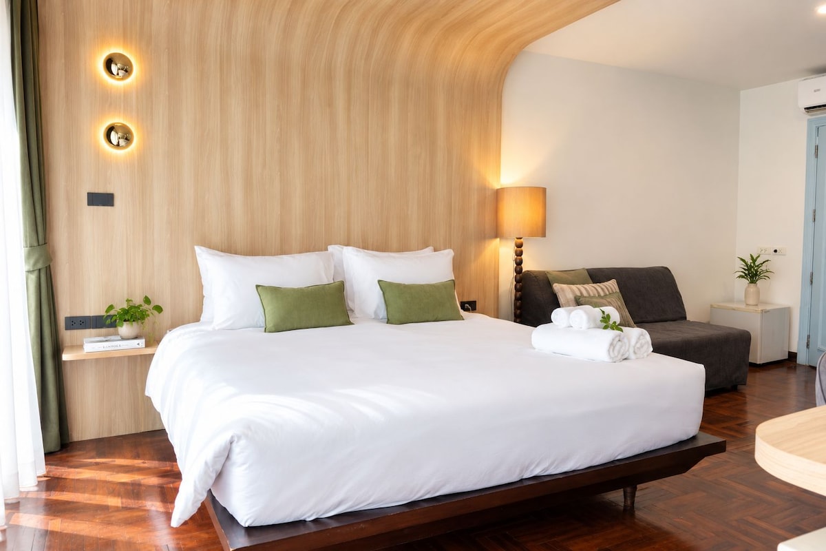 A hotel room with a large bed, bedside table, lamp, and a small sofa.