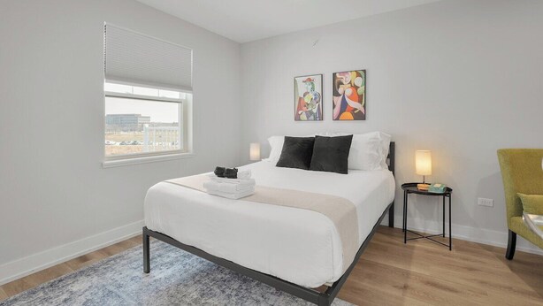 Ein Hotelzimmer mit Bett, Fenster, Wandbild, Nachttisch mit Lampe und Sessel.