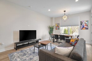 A modern living room with a flat-screen TV, a sofa, a coffee table, and a dining area with chairs and a table. - Luxe Warrenville 4BR | Patio + Pickleball Court (Warrenville)