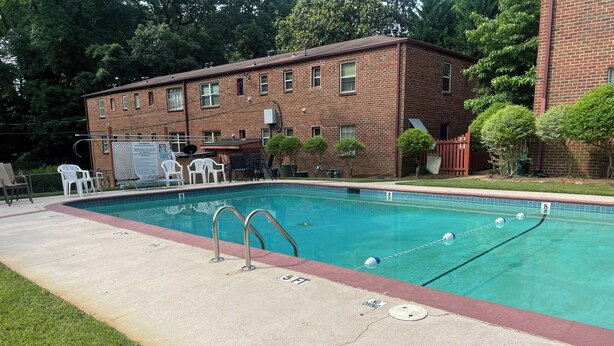 Ein rechteckiger Pool mit roter Umrandung, zwei Metallleiter und eine Poolterrasse mit weißen Stühlen und einem Tisch.