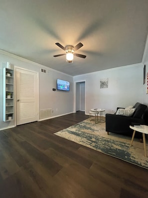 A living room with a ceiling fan, a flat-screen TV mounted on the wall, a black sofa, a small round table, and a patterned rug. - Renovated Condo: Your home away from home (Decatur)