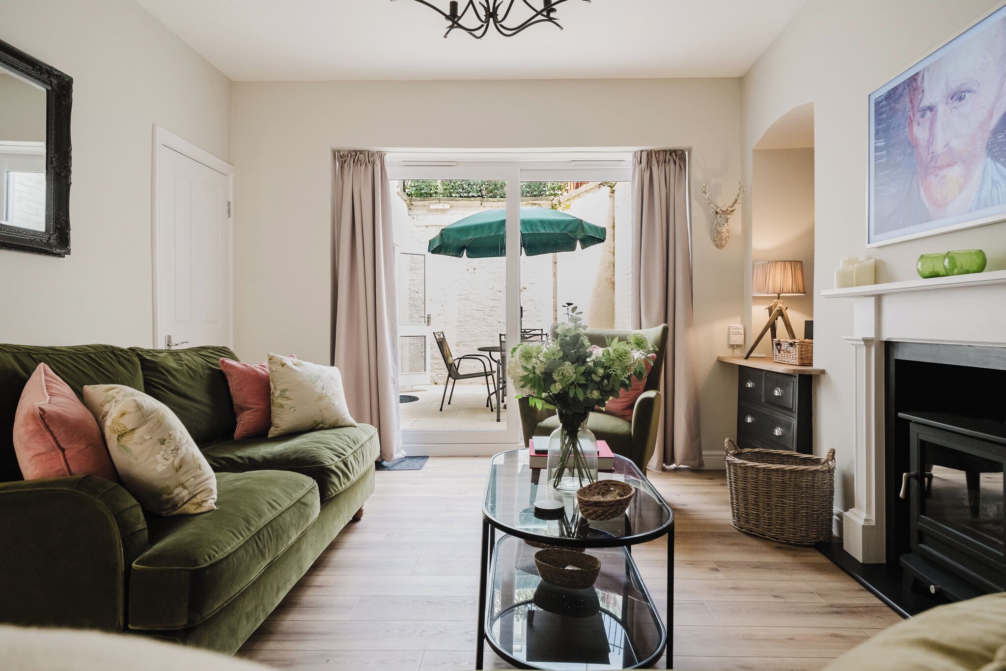 A living room with a green sofa, a glass coffee table, a fireplace, and a view through a glass door to an outdoor area with a green umbrella.