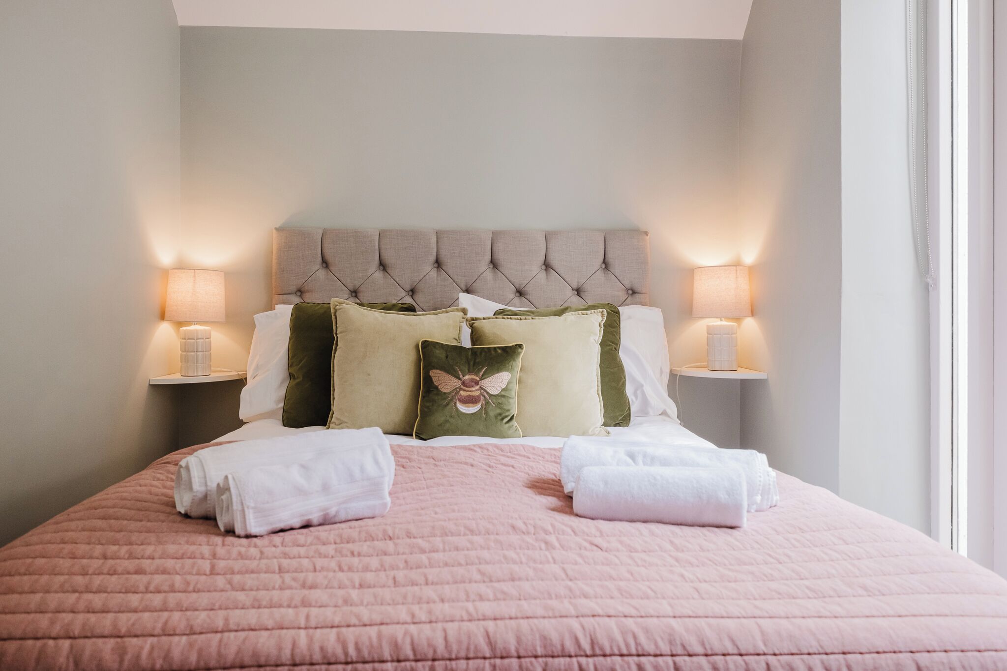 Una cama bien hecha con colcha rosa, cabecera capitonada y dos lámparas de noche.