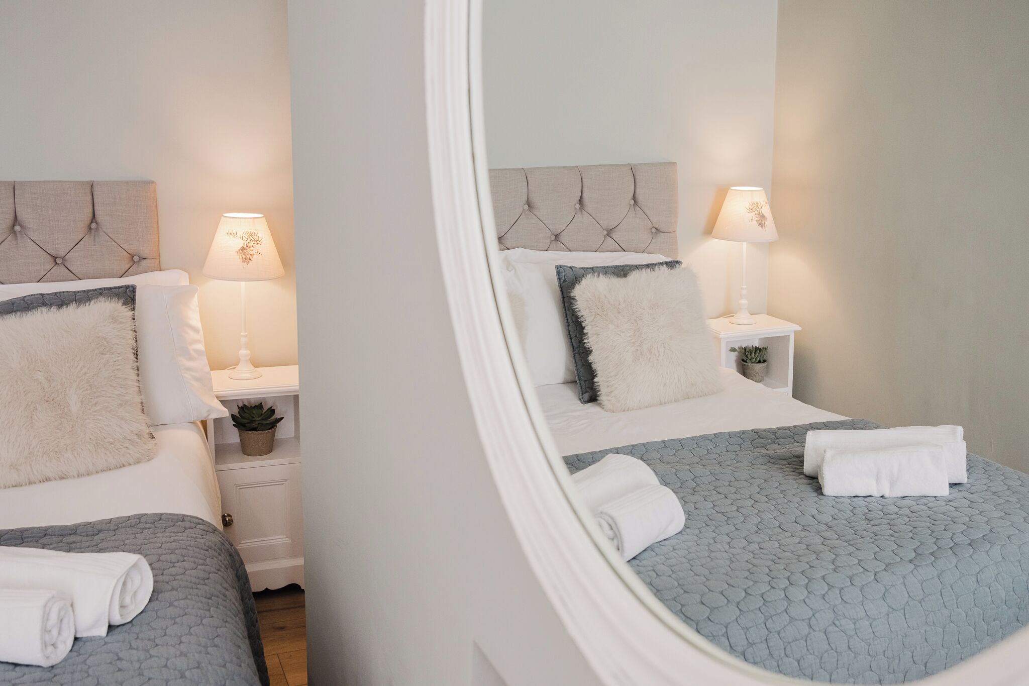 A neatly made bed with a grey textured bedspread, two white pillows, and a fluffy white throw. There is a bedside table with a lamp and a small potted plant.
