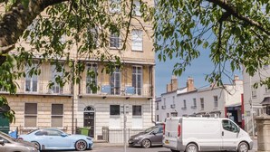 A street view with a row of residential buildings, parked cars, and a tree providing shade. - Stunning Flat in Heart of Cheltenham (Cheltenham)