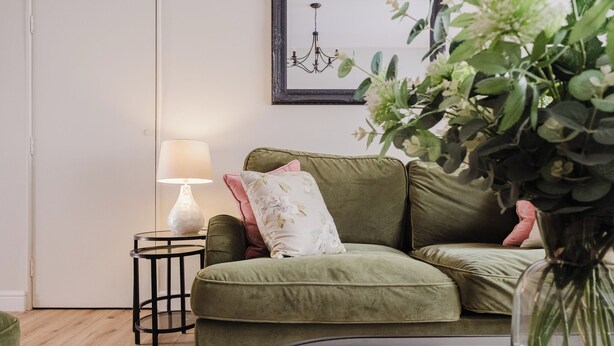 A green sofa with floral cushions, a round side table with a lamp, and a framed picture on the wall. - Stunning Flat in Heart of Cheltenham (Cheltenham)
