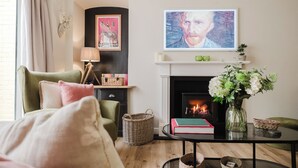 A cozy living room with a fireplace, a green armchair, a coffee table with books and a vase of flowers, and a painting above the mantel. - Stunning Flat in Heart of Cheltenham (Cheltenham)
