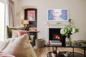 Ein gemütliches Wohnzimmer mit Kamin, einem grünen Sessel, einem Couchtisch mit Büchern und einer Blumenvase sowie einem Gemälde über dem Kamin.