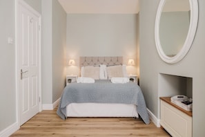 A bedroom with a bed, bedside lamps, a mirror, and a small wooden side table. - Stunning Flat in Heart of Cheltenham (Cheltenham)
