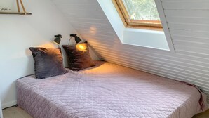 Ein Einzelbett mit gesteppter Bettdecke, zwei Wandleuchten und ein Oberlicht in einem Raum mit weißen Wänden und Holzbalken.